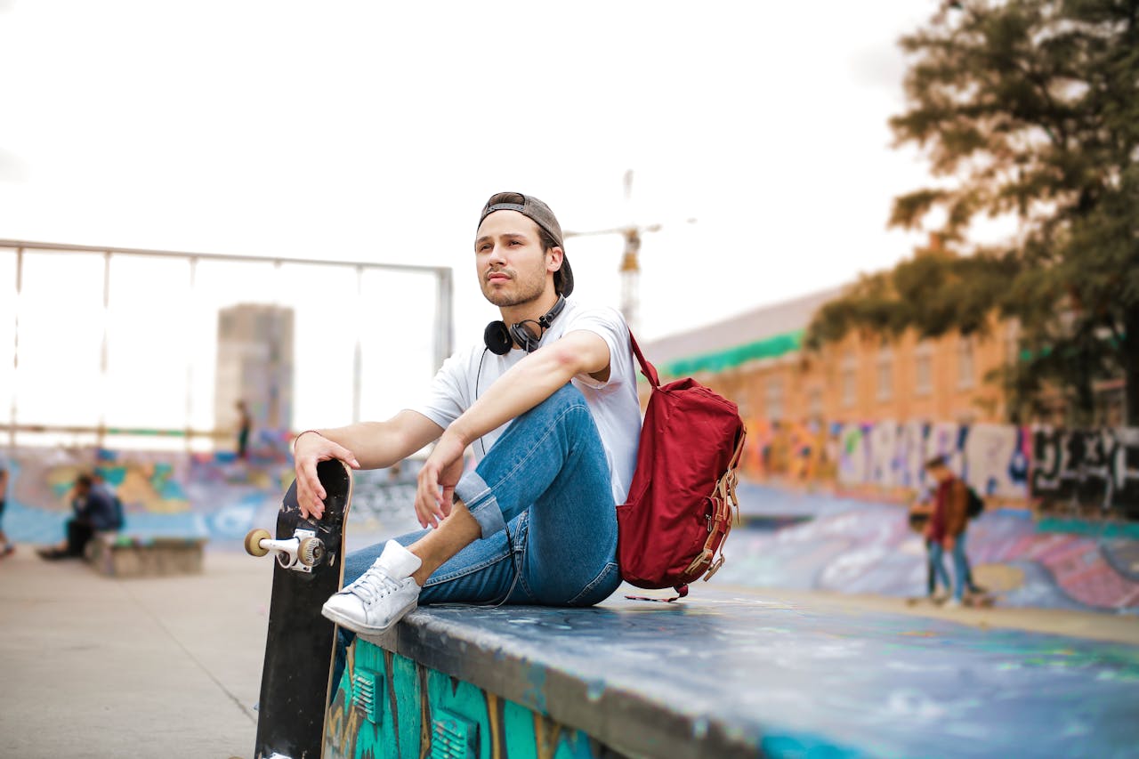 why-choose-us Man in casual outfit with longboard rucksack and headphones on neck sitting on ramp in skatepark and looking away